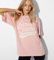A woman with blonde hair posing for the camera. She is wearing black leggings, and a pale pink t-shirt with graphic text 'Lona Jane Sports Club' on it. She is holding the collar of her shirt in a posed position.
