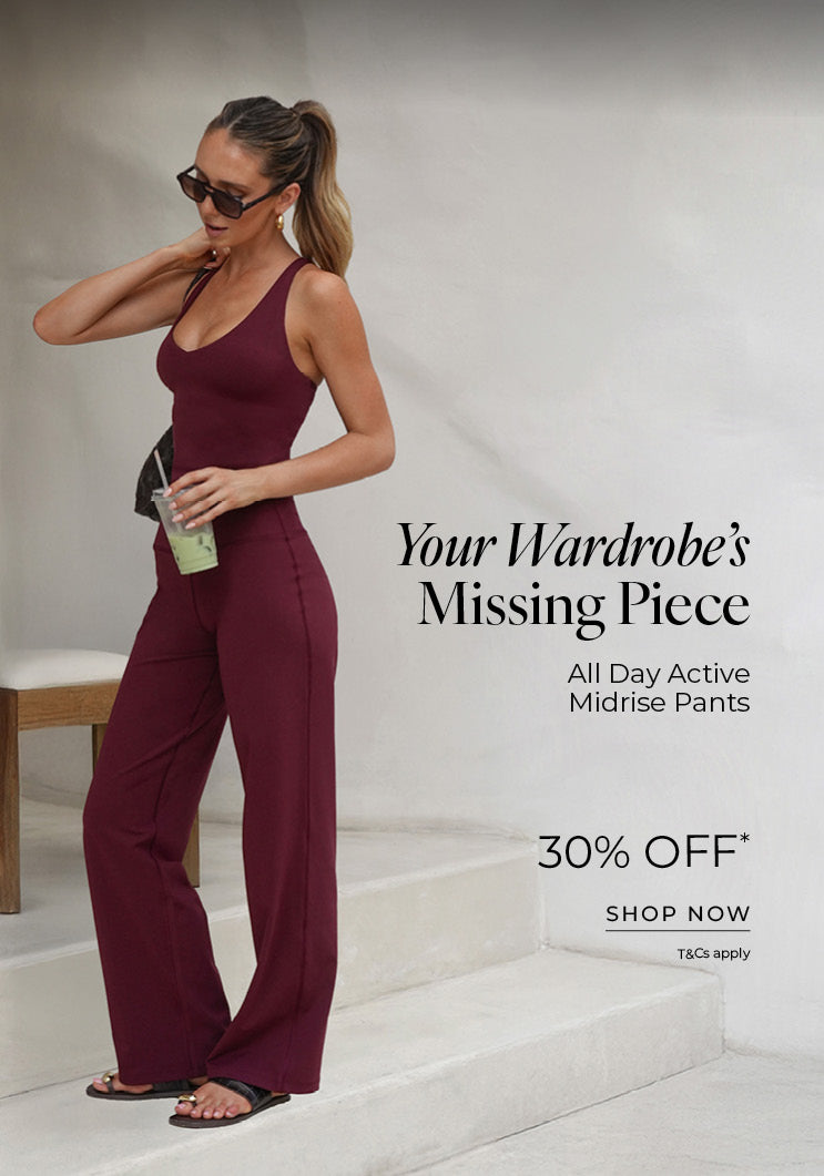 A woman stands on outdoor steps wearing a burgundy fitted tank top and matching wide-leg active pants, accessorized with sunglasses and holding a drink. Text: Your Wardrobe's Missing Piece. All Day Active Midrise Pants 30% Off* T&Cs apply - Shop Now