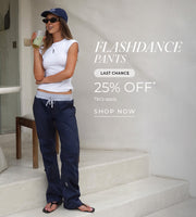 A woman stands on light stone steps holding a takeaway drink, wearing a white sleeveless top, navy Flashdance pants with a drawstring waist, a navy baseball cap, sunglasses, and sandals. Text: Flashdance Pants Last Chance 25% Off* T&Cs Apply. Shop Now