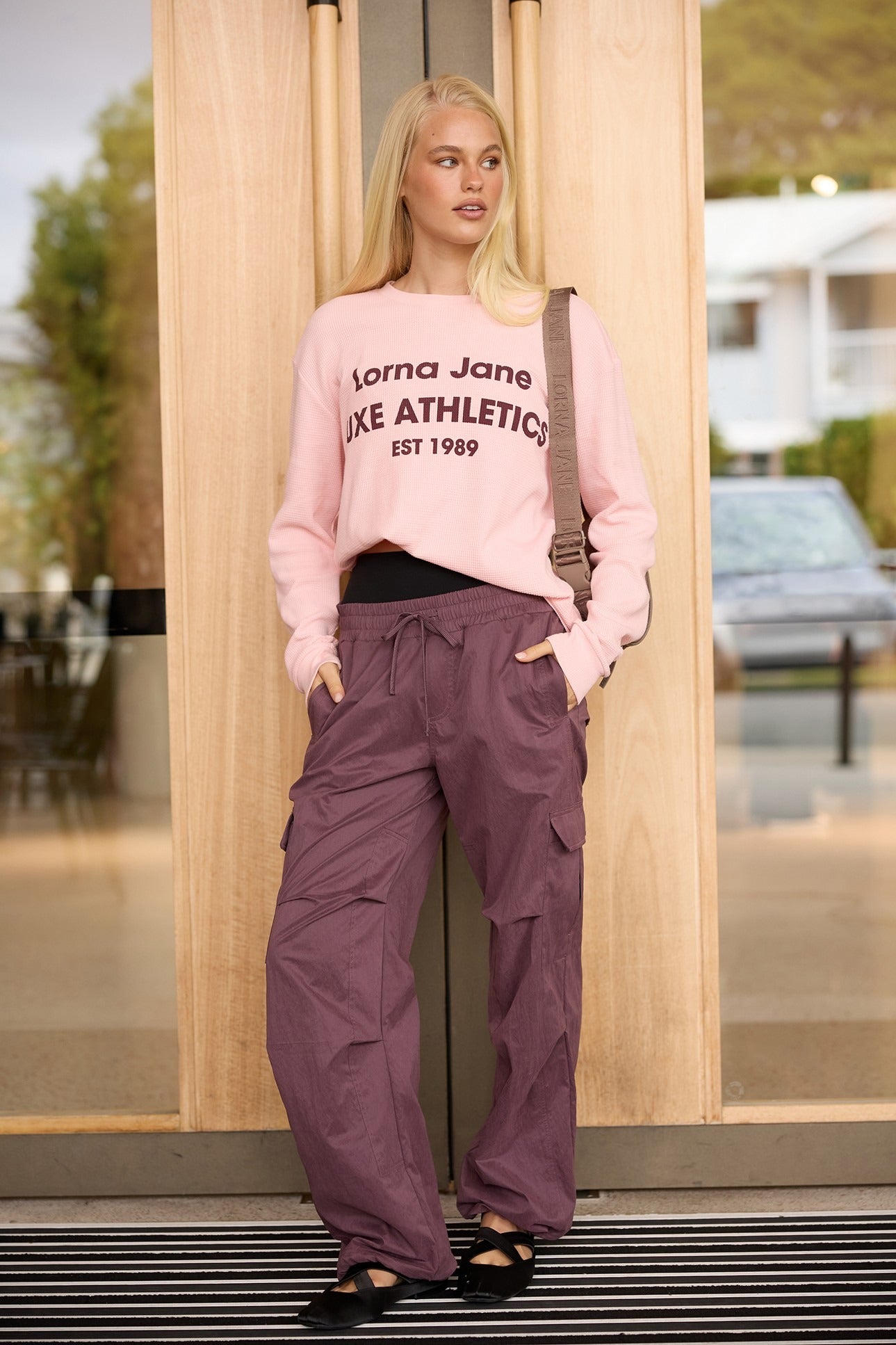 Blonde woman in a pink waffle-knit graphic sweatshirt and mauve cargo pants with a crossbody bag, standing in a doorway.