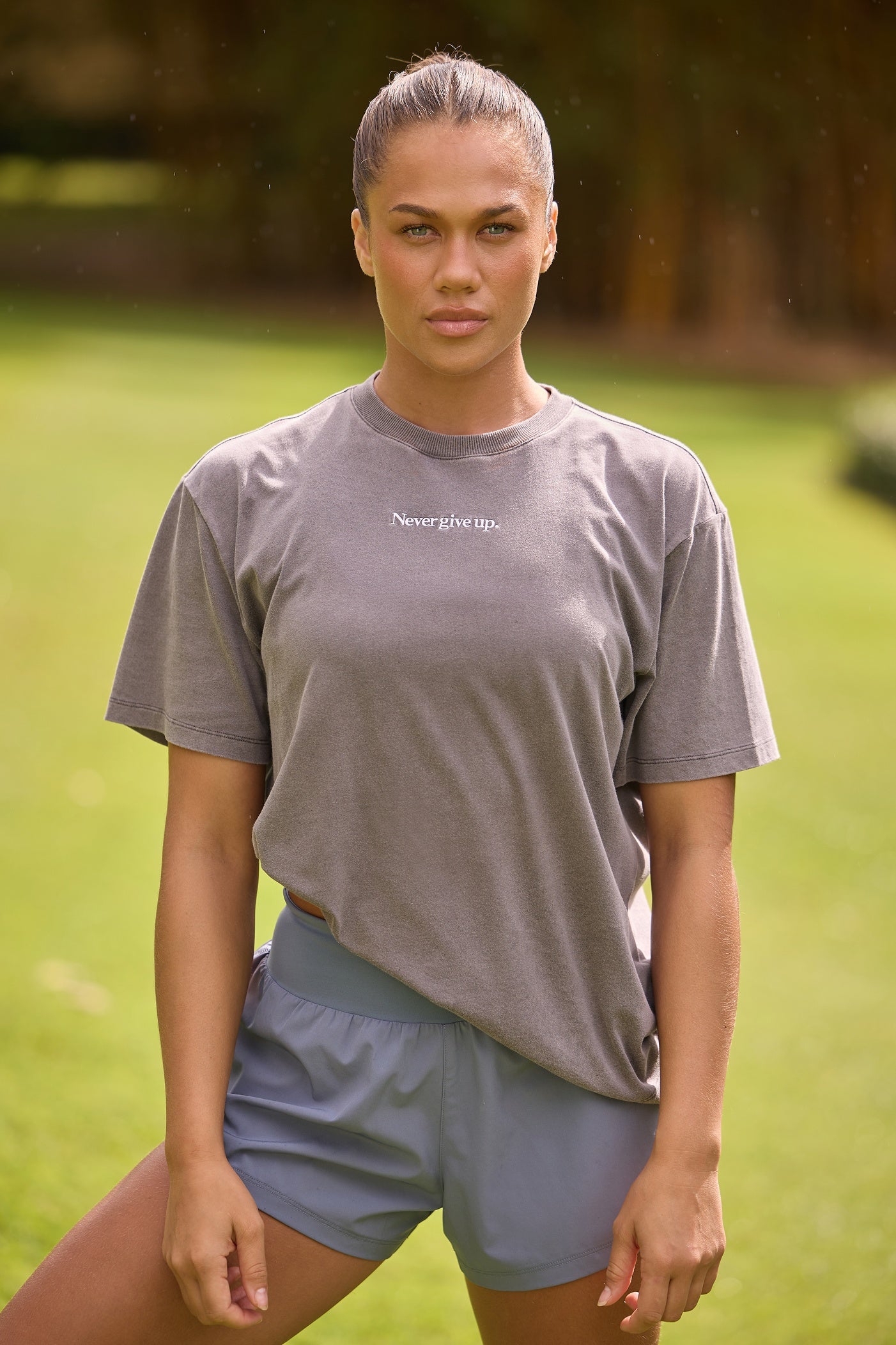 A woman stands outdoors on a grassy field wearing a grey oversized T-shirt with the text 
