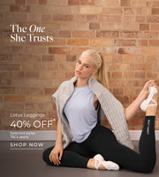 A woman seated on a studio floor in a stretching pose against a brick wall. She wears a light blue sleeveless top, black leggings, white ballet-style shoes, and a grey cable-knit sweater draped over her shoulders. Text: The One She Trusts. Lotus Leggings 40% Off* Selected styles. T&Cs Apply - Shop Now