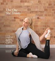 A woman seated on a studio floor in a stretching pose against a brick wall. She wears a light blue sleeveless top, black leggings, white ballet-style shoes, and a grey cable-knit sweater draped over her shoulders. Text: The One She Trusts. Lotus Leggings 40% Off* Selected styles. T&Cs Apply - Shop Now