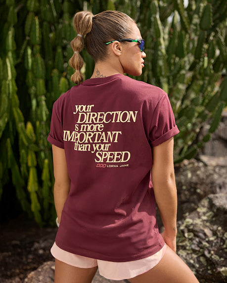 A woman stands outdoors with their back turned, wearing a burgundy oversized T-shirt featuring the statement, 