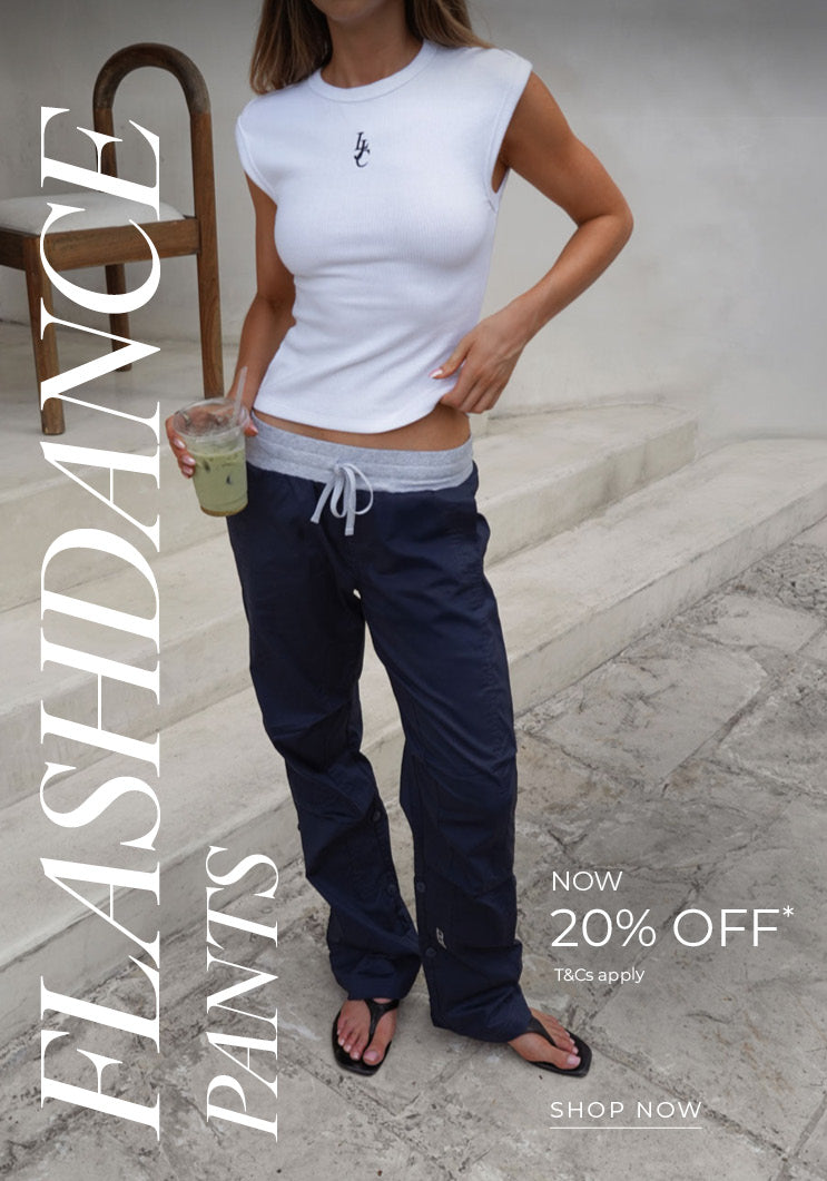 Woman wears navy Flashdance Pants with a grey drawstring waistband, styled with a white sleeveless top, sunglasses, and a navy cap, holding an iced drink in a bright, sunlit stone setting. Text: Flashdance Pants Now 20% Off* T&Cs apply - Shop Now