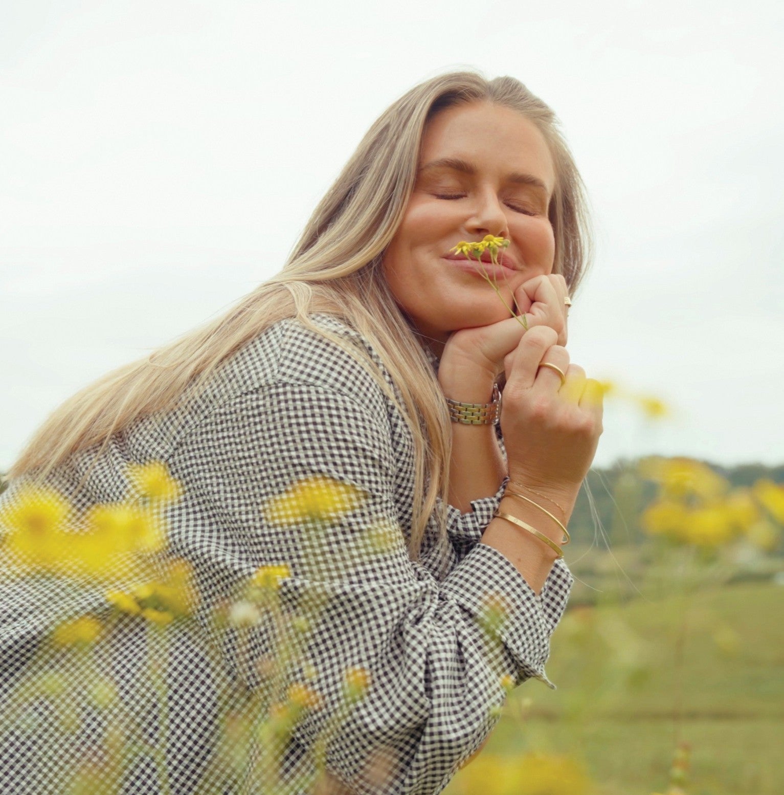 A blonde woman with her eyes closed smiles softly as she holds a small yellow flower to her nose, resting her chin on her clasped hands. She wears a black-and-white gingham shirt with gold bracelets and rings, framed by blurred yellow wildflowers in a green field.