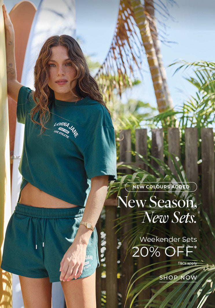 Woman in a deep teal Lorna Jane cropped tee and matching shorts, standing beside surfboards in a tropical garden. Text: New Colours Added. New Season, New Sets. Weekender Sets 20% Off* T&Cs apply - Shop Now