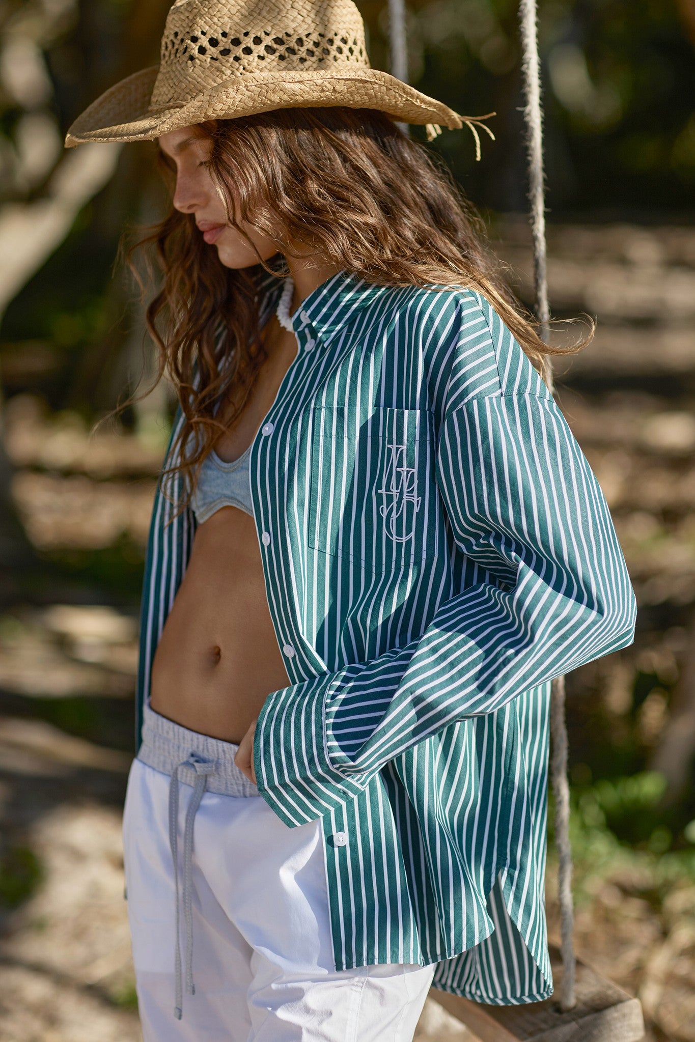 Woman in a straw hat and green-and-white striped oversized shirt with LJ monogram, worn open over a denim bralette with white drawstring pants, standing outdoors in dappled sunlight.