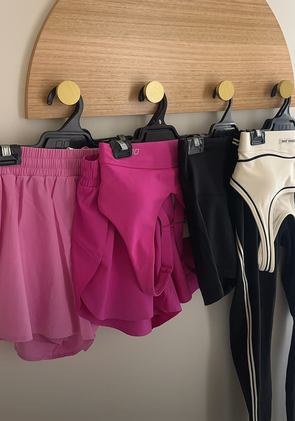 Four Lorna Jane bottoms hung on clip hangers from a wooden peg rail, a light pink flowy skirt, magenta shorts, black bike shorts, and cream-and-black panelled leggings.