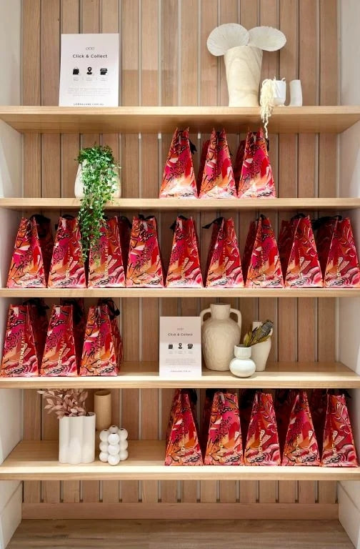 Wooden retail shelving display featuring multiple rows of colorful red and pink patterned Click & Collect bags, styled with decorative ceramic vases, plants, and signage in a modern store interior.