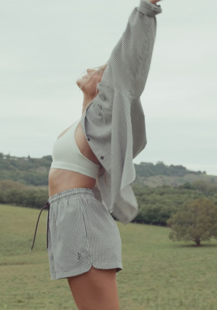 A woman moves through a rolling green valley and lush tropical garden in three elevated warm weather looks, a white crop top with gingham shorts and an oversized striped shirt, a white silk slip dress, and a textured white knit top with striped shorts. Lorna Jane lifestyle activewear.