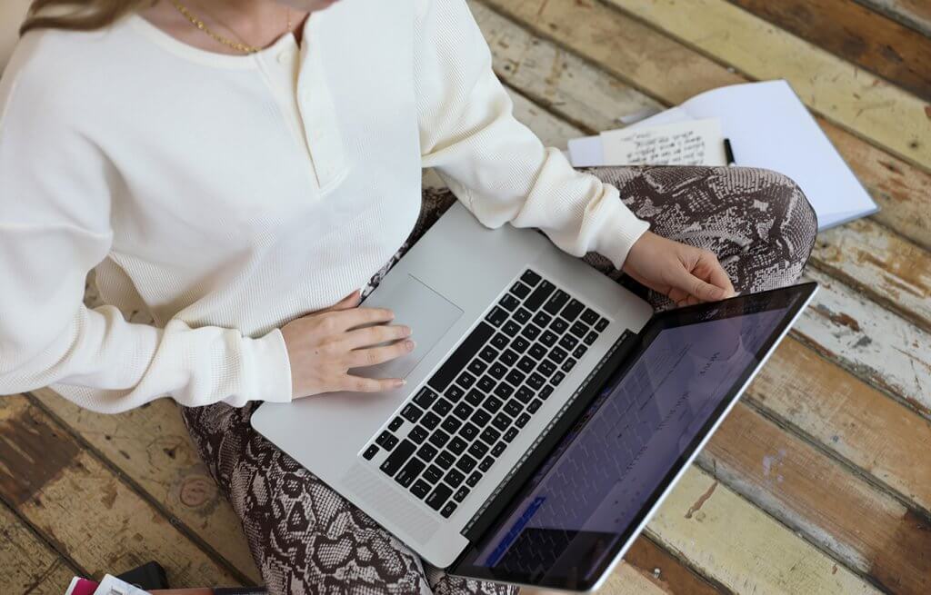 lorna jane sitting cross legged on floor holding laptop wearing cream knitted top and snake print leggings
